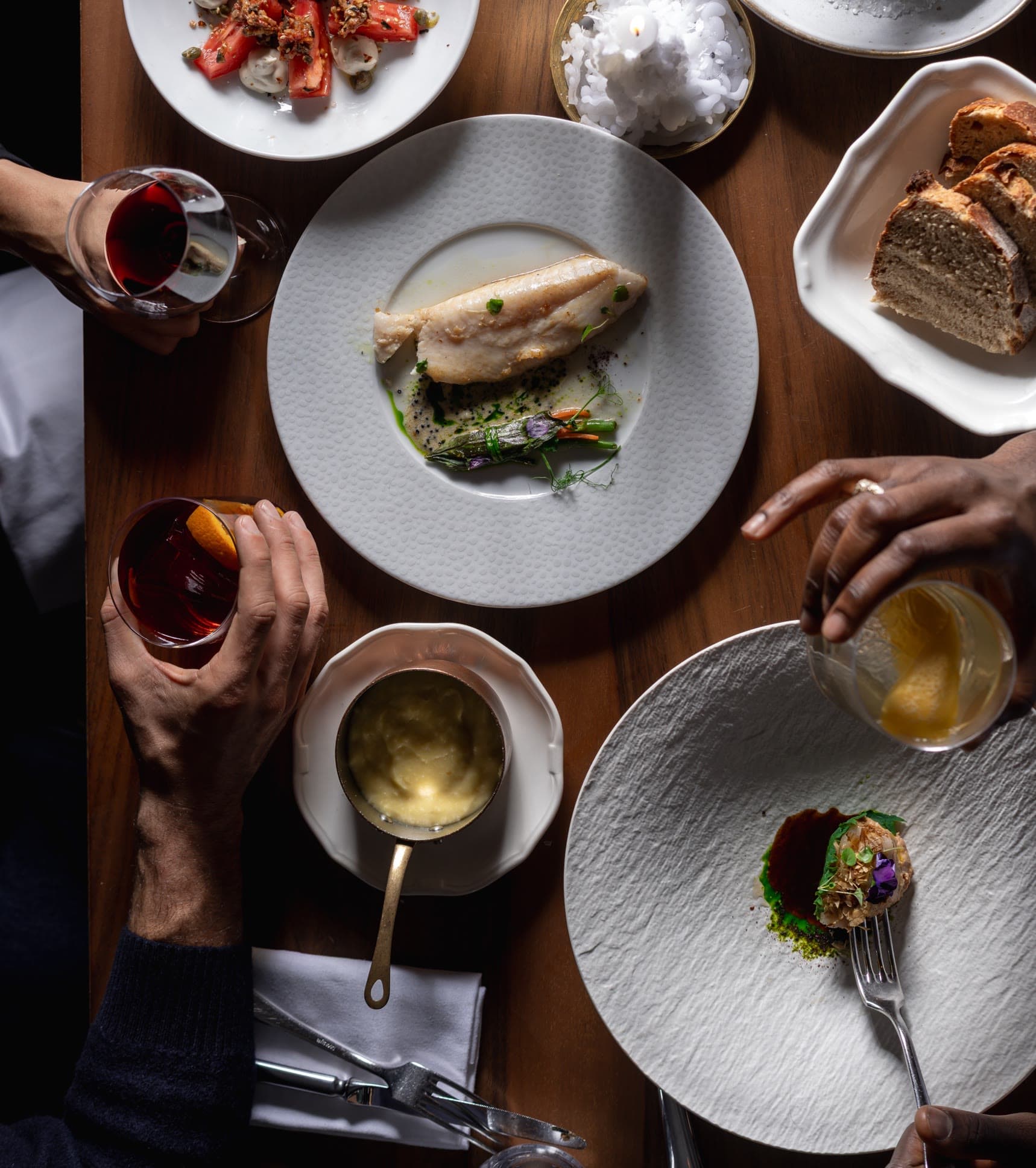 A shared table seen from above: fish, tomatoes, purée, bread and glasses of wine in the guests' hands